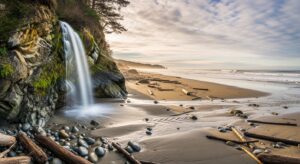 A scenic view of Alamere Falls cascading down a rocky cliff into the ocean at a beach, with logs and pebbles scattered along the shoreline under a cloudy sky